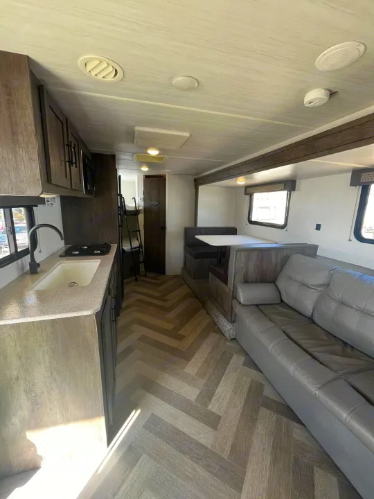 Bright RV rental interior showing a kitchen (left), dinette, and sofa (right). Herringbone floors, bunk beds visible. Ready for travel.. Forest River EVO 2021