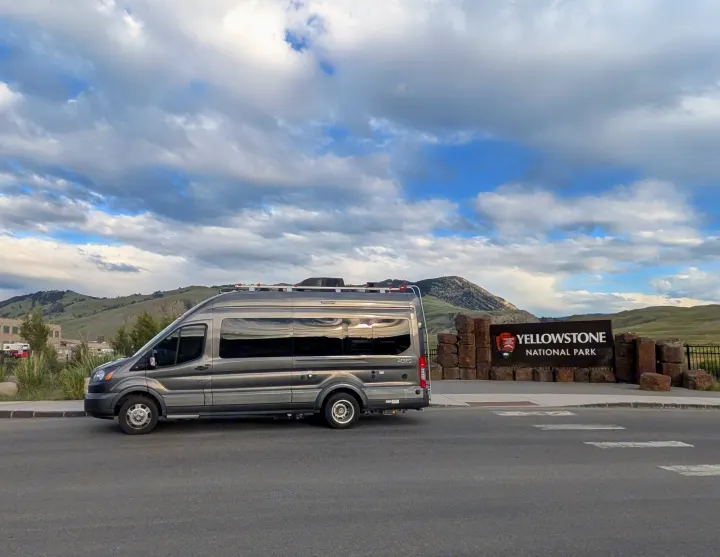 Gray RV rental van at the iconic Yellowstone National Park sign, with mountains in the background under a cloudy sky.. Winnebago Paseo 2017