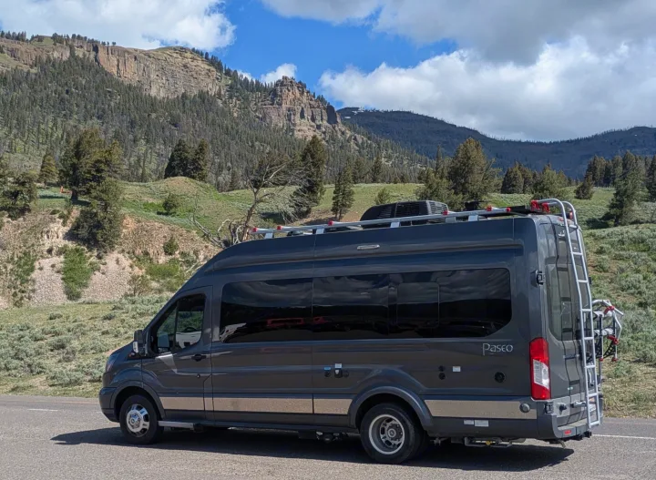 A dark gray RV rental with a bike rack is parked on a scenic mountain road, surrounded by trees under a cloudy sky.. Winnebago Paseo 2017