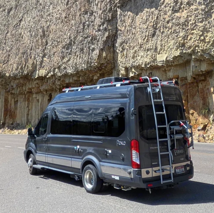 Dark gray Winnebago Paseo RV rental with roof rack & bike carrier, parked next to a textured rock cliff.. Winnebago Paseo 2017