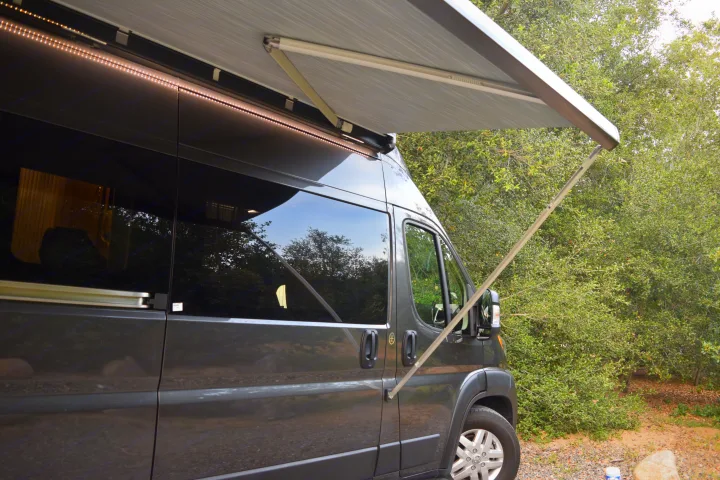 Side view of a grey RV rental with its awning extended and bright LED lights on, parked amidst green trees.. Thor Sequnce 2023