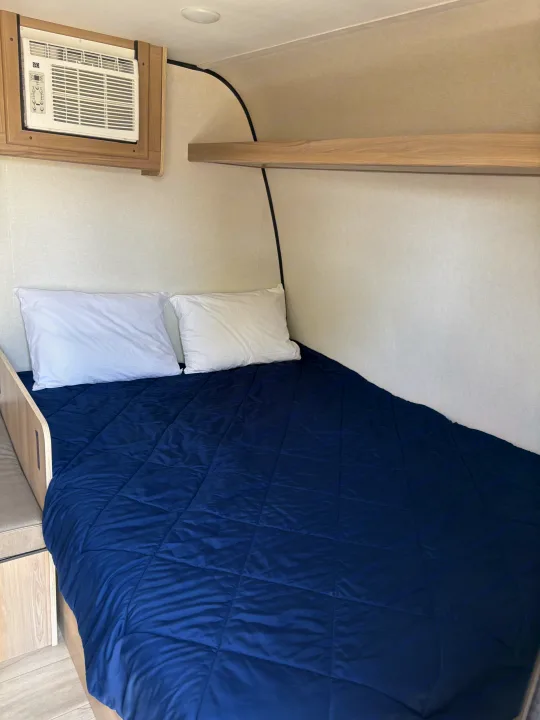Cozy RV rental bedroom with dark blue blanket, white pillows, AC unit, and storage shelf.. Keystone RV Coleman 2025