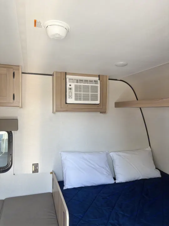 Inside an RV rental: a cozy bed, window AC unit, and overhead storage. Smoke detector visible on ceiling.. Keystone RV Coleman 2025