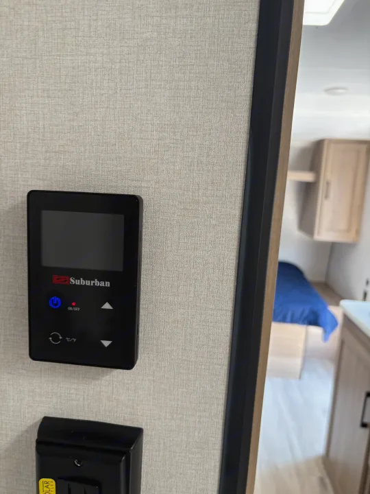 Suburban control panel on an RV rental wall, managing climate. View into the bedroom with a blue bed and light cabinets.. Keystone RV Coleman 2025