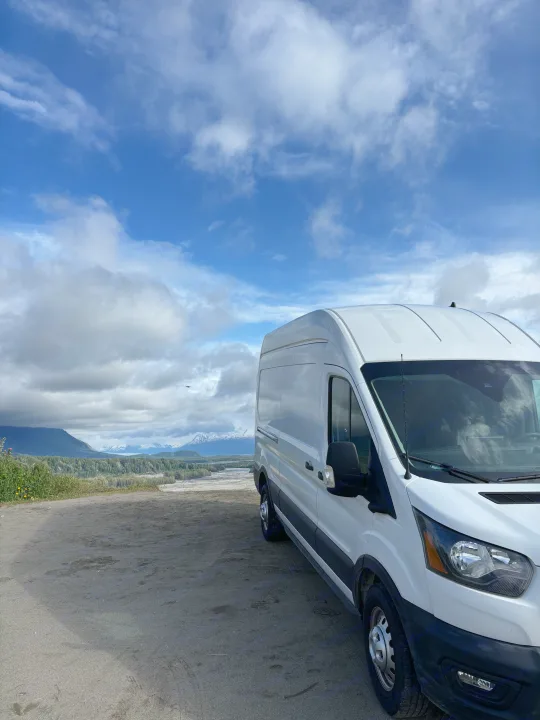 White RV rental van overlooks vast Alaskan landscape: mountains, river, and forests under cloudy sky.. Ford Transit 2022