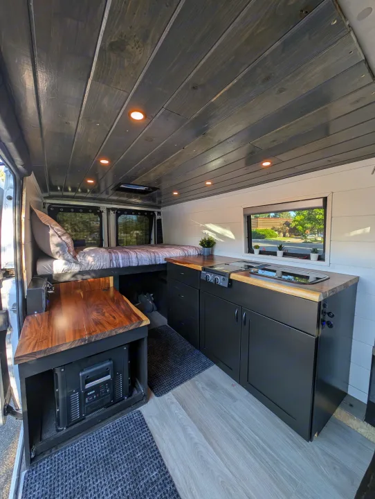 Modern camper van interior with a bed, kitchen, wood counters, and dark cabinets. Ideal for an rv rental adventure.. Custom Ram Promaster 1500 2017