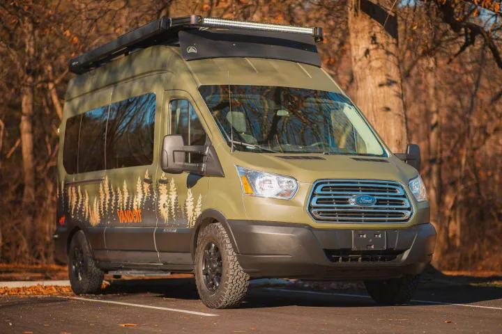 An olive green Ford Transit camper van with custom tree graphics, off-road tires, and a roof rack, ready for your next RV rental trip.. Vandoit LIV 2019
