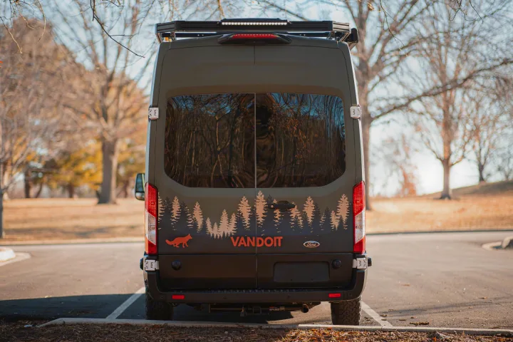 Rear view of a dark green VANDOIT custom rv rental van with pine tree & fox decals, parked in an outdoor setting.. Vandoit LIV 2019