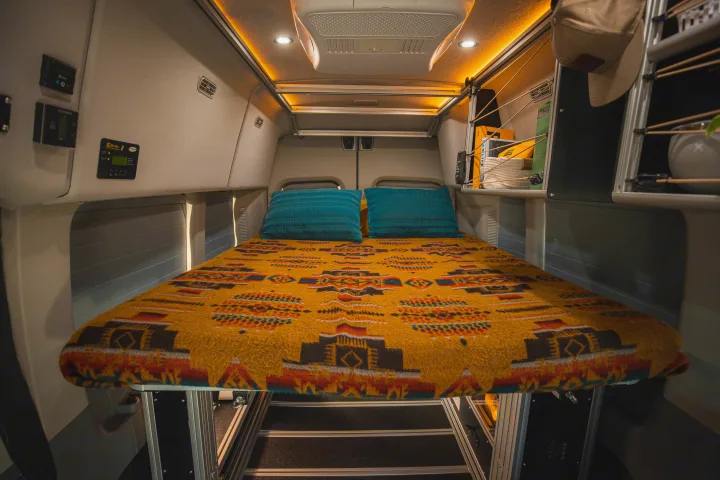 Cozy RV rental interior with a vibrant patterned bed, overhead lighting, and built-in storage, perfect for van life adventures.. Vandoit LIV 2019