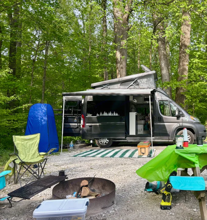 Serene forest camp setup featuring an RV rental with pop-top, awning, fire pit, and picnic area.. Thor Motor Coach 18A Rize 2023