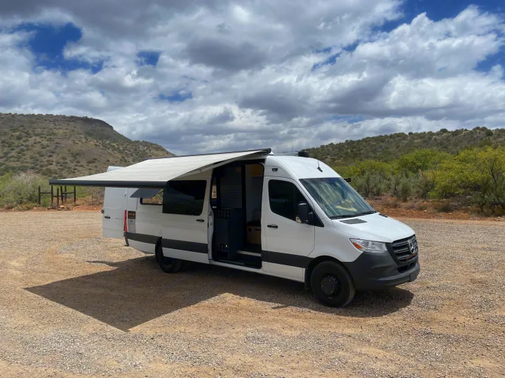 White camper van with awning extended, parked on a gravel lot amidst arid hills. Ideal for your next RV rental outdoor adventure.. Mercedes Sprinter 2023