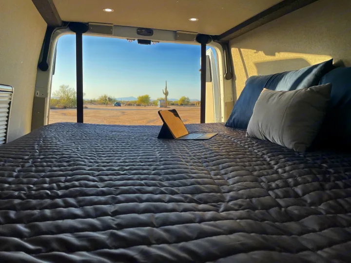 RV rental: Relax in a comfy bed in your camper van, enjoying desert views with saguaro cacti. Laptop on bed for convenience.. Mercedes Sprinter 2023