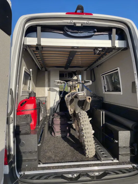 This **RV rental** camper van interior features a dirt bike, elevated bed, and ample storage for adventure gear.. Mercedes Sprinter 2023