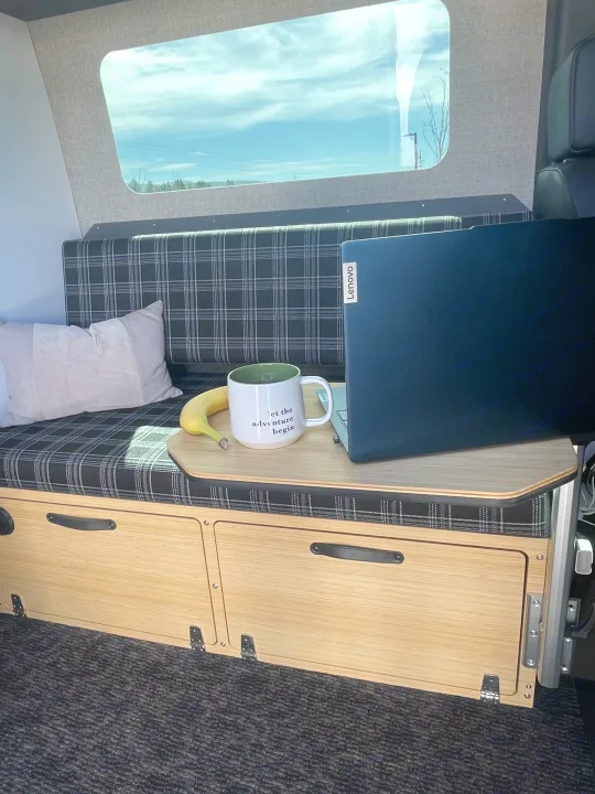 RV rental: Plaid bench with a pillow, wooden table holding a Lenovo laptop, an "adventure" mug, and a banana. Perfect mobile workspace.. Mercedes Sprinter 2023