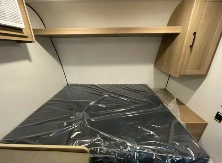 Compact RV rental sleeping area features a new, plastic-wrapped mattress, shelf, and cabinet overhead.. Other Other 2025