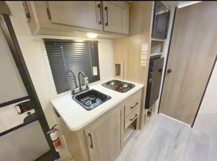 An RV rental's compact kitchen. Features include a sink, stovetop, microwave, and refrigerator, set in a modern, space-saving design.. Other Other 2025
