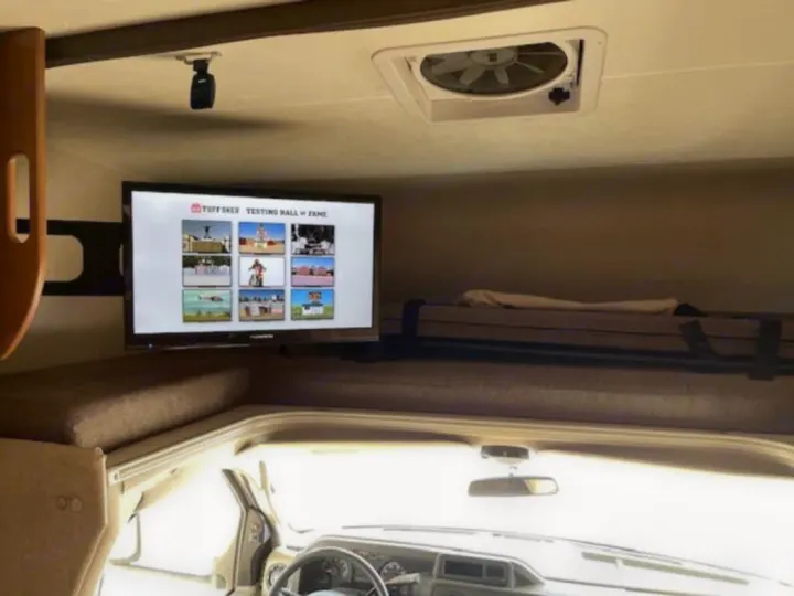 Inside an RV rental, a TV shows 'Tuff Shed' content. An overhead bunk & driver's cab are visible. Perfect for adventure!. Coachmen Leprechaun 2019