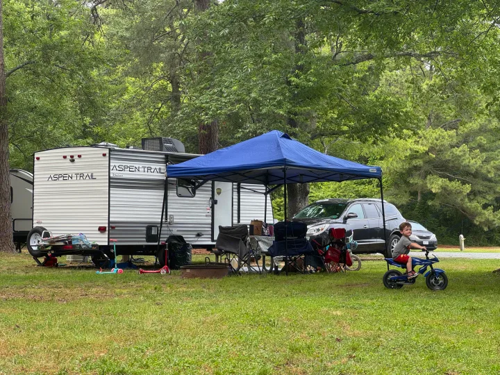 RV rental at a family campground. A child rides a toy motorcycle while camping beside a travel trailer and blue canopy.. Dutchman Aspen Trail 2025