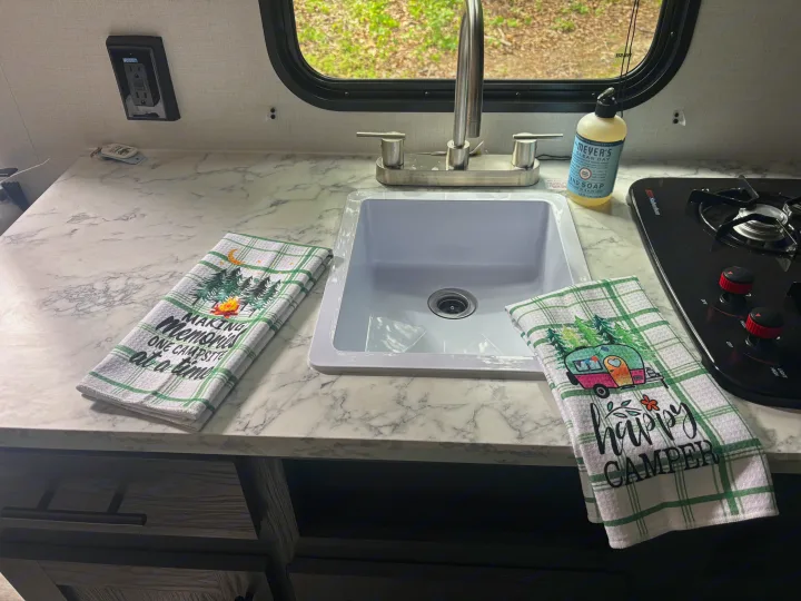 RV rental interior features a functional kitchen with a sink, faucet, stove, and charming camping towels on the counter.. Dutchman Aspen Trail 2025