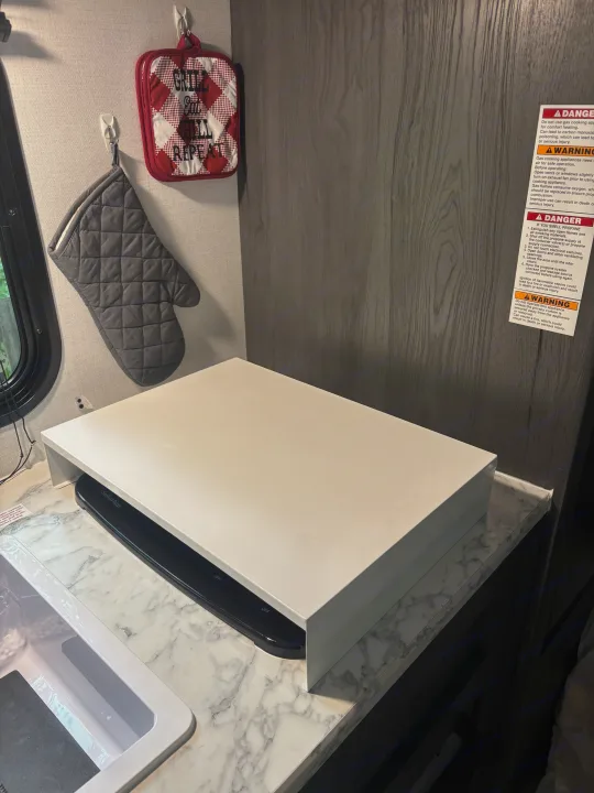 This RV rental kitchen features a white cover over the stovetop, a sink, and "Grill Chill Repeat" potholders. Safety warnings are visible on the wall.. Dutchman Aspen Trail 2025