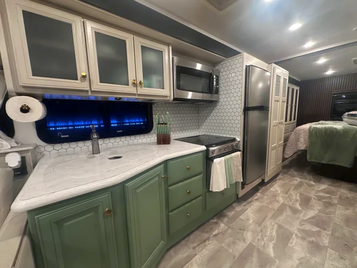 Modern RV rental kitchen with green/white cabinets, stainless steel appliances, hexagon tile backsplash, and a bedroom visible.. Fleetwood Flair 2021