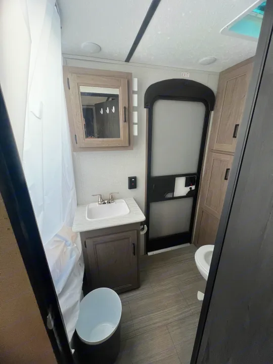 Explore this RV rental's compact bathroom, featuring a sink, vanity, mirror, shower with white curtain, toilet, and storage.. Keystone RV Hideout 2021