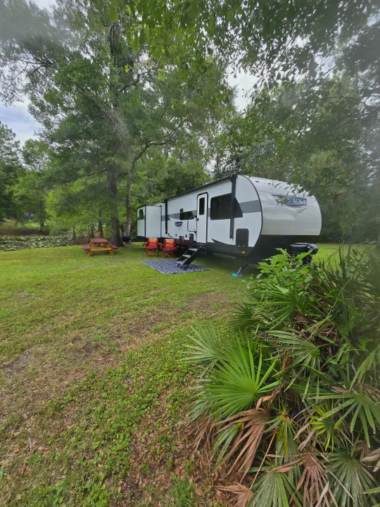Serene RV rental campsite: a white travel trailer by a pond with a picnic table & chairs under lush trees. Perfect outdoor getaway.. Forest River Salem Platinum 2024