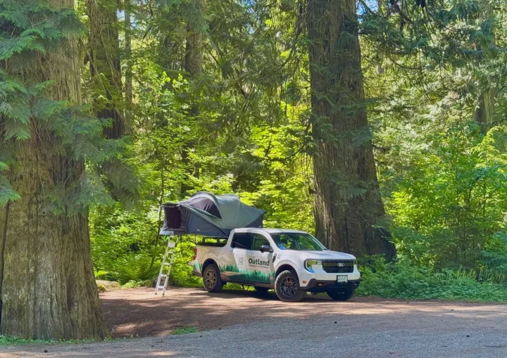 White Outland adventure RV rental truck with deployed rooftop tent in a lush, sun-dappled forest. Ideal for nature camping.. Ford Maverick 2025