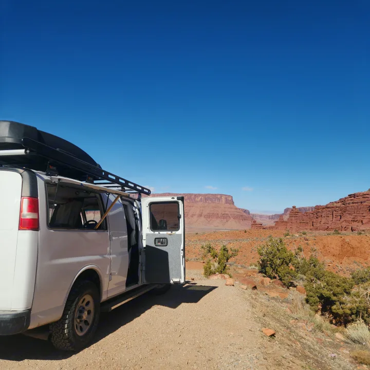 Scenic rv rental van adventure: white van with open door on a desert road, majestic red cliffs under clear sky.. Chevrolet Express 1500 AWD 2014