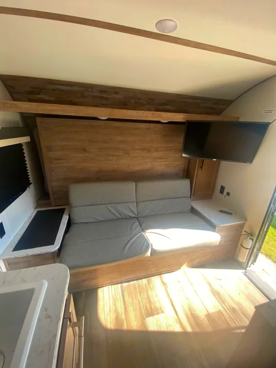 Inside this modern RV rental: light gray couch, mounted TV, wood-look interior, and small kitchen sink area.. Gulf Stream Grand River 2024