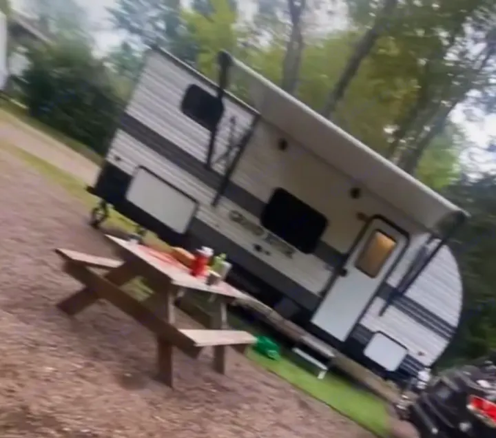 An RV rental travel trailer is parked at a gravel campsite next to a picnic table with food items. Enjoy the outdoors!. Gulf Stream Grand River 2024