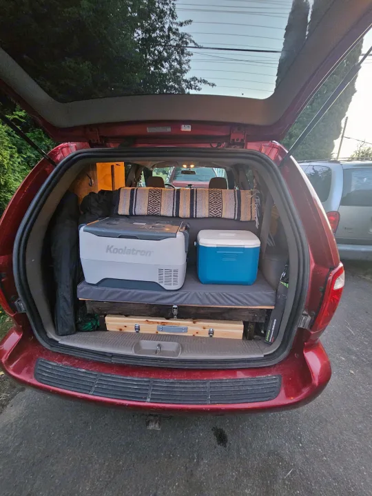 Rear of a red minivan, converted for car camping with a sleeping platform, portable fridge, and cooler. A smart RV rental alternative.. Dodge Caravan 2003
