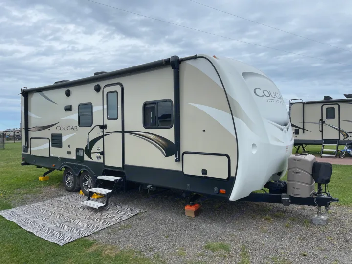 A tan and white travel trailer RV rental, featuring an outdoor mat and steps, parked on gravel in a campsite under a cloudy sky.. Keystone RV Cougar 2018