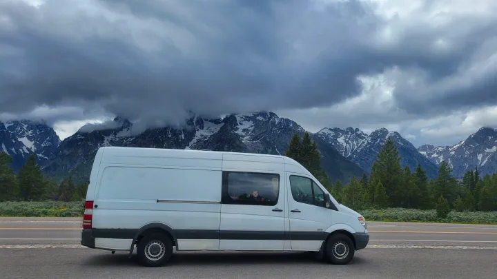 White RV rental van stopped roadside with child inside, facing dramatic snow-capped mountains under cloudy sky.. Freightliner Sprinter 2011
