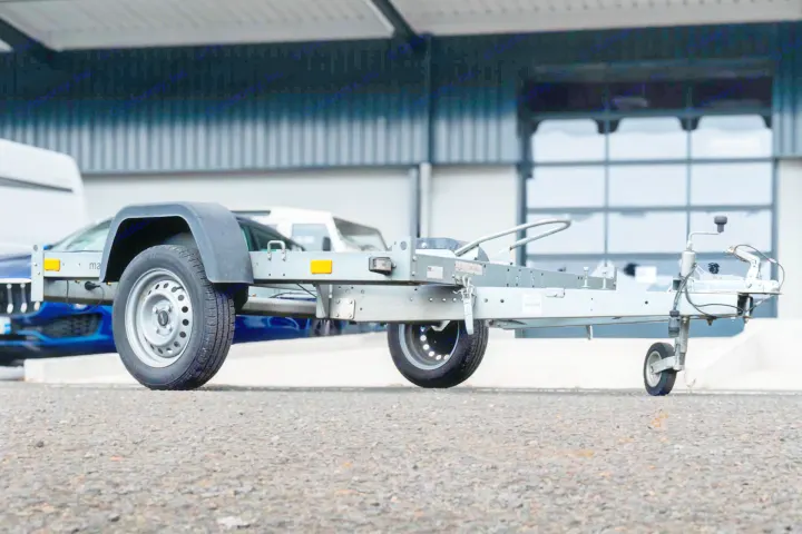 A light gray utility trailer, ideal for an RV rental trip or hauling, parked on asphalt with vehicles and a building in the background.. RemorK Porte Moto 2025