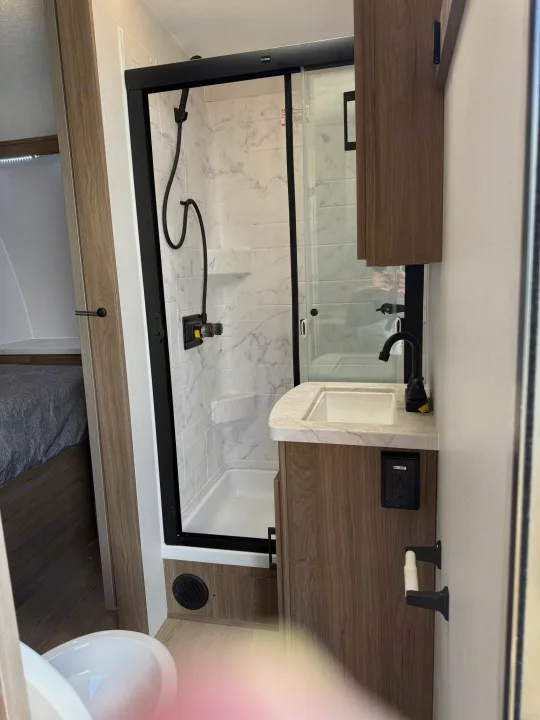Modern RV rental bathroom: compact shower, white sink, wood vanity, black fixtures. Partial toilet view.. Coachmen Adrenaline 2025