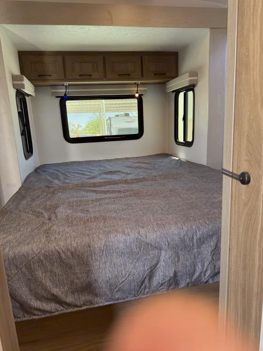 Inside an **rv rental** bedroom: a large bed with gray cover, overhead cabinets, and windows with blinds. Ready for adventure.. Coachmen Adrenaline 2025