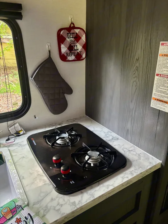 Inside an RV rental, a compact kitchen features a black 2-burner gas cooktop, marble-look countertop, sink, and hanging mitt/potholder.. Dutchman Aspen Trail 2025