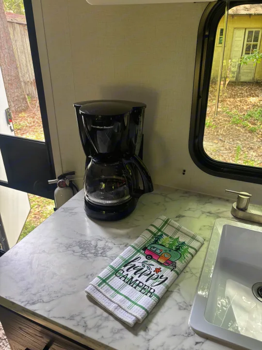 An RV rental's kitchen counter with a coffee maker, sink, and "Happy Camper" towel.. Dutchman Aspen Trail 2025