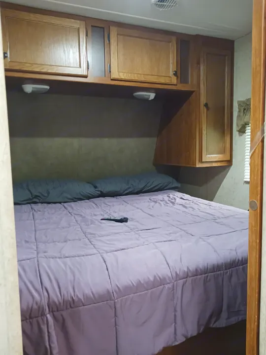 Comfy RV rental bedroom features a purple comforter, pillows, remote, and overhead wooden cabinets for your travel comfort.. Heartland RVs Trail Runner 2013