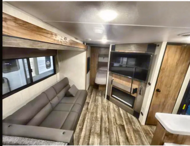 Modern RV rental interior featuring a living area with a grey sofa, large TV, fireplace, and bed visible beyond.. Forest River Campsite Reserve 26CJ 2025
