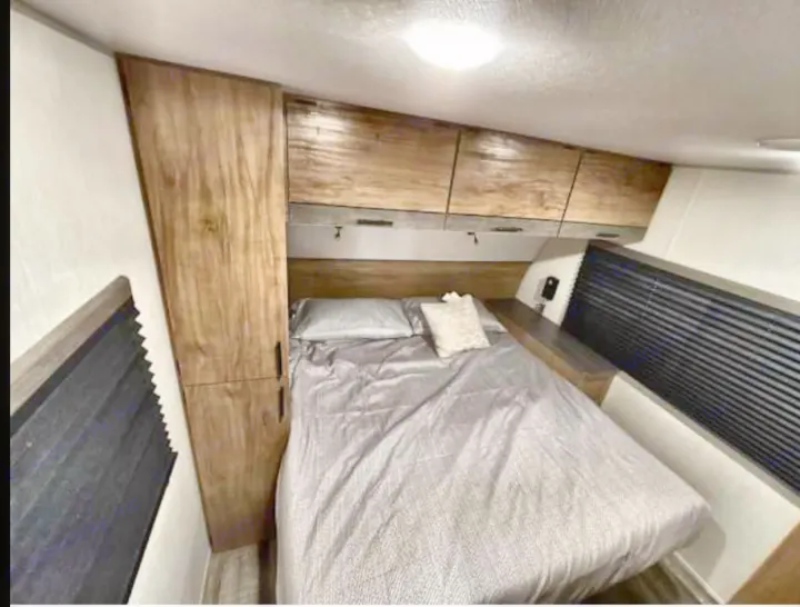 RV rental bedroom: A comfortable bed with grey bedding, wood-tone cabinets, and window blinds.. Forest River Campsite Reserve 26CJ 2025