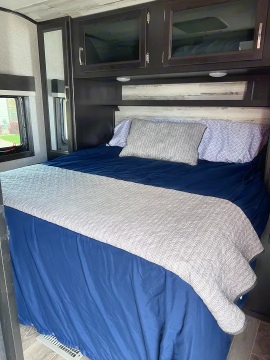 This comfortable RV rental bedroom features a blue bed with a gray quilt, dark cabinets, and windows.. Jayco Jay Feather 2021