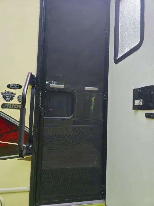 Side view of an RV rental entrance, featuring a black screen door, a sturdy grab handle, and the motorhome's light exterior.. Jayco Redhawk 2017
