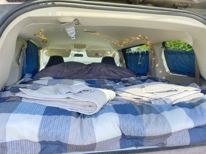 Inside an RV rental setup: a cozy bed with plaid blankets, towels, and string lights create a comfy car camping retreat.. Dodge Grand Caravan 2008