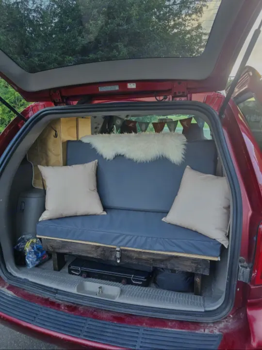 Red minivan's tailgate open, revealing a DIY camper conversion with a blue cushioned bench bed. Ideal for RV rental travel.. Dodge Caravan 2006