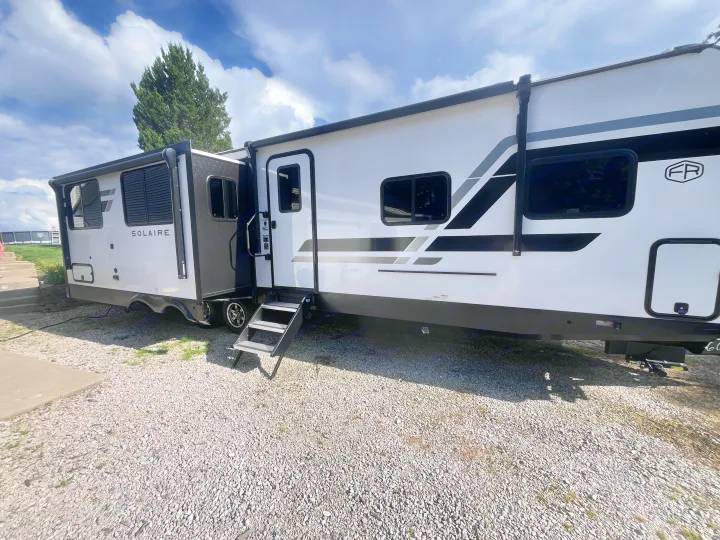 White & black Solaire RV rental parked on gravel at a sunny campsite with sky & tree.. Forest river 306RKTS Palomino 2025