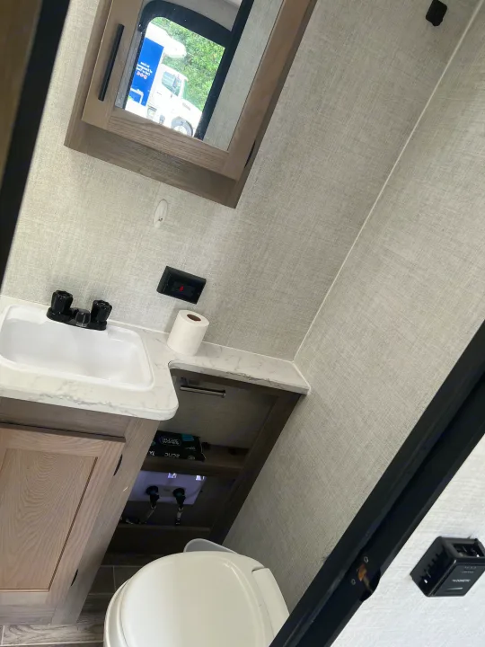 An RV rental bathroom featuring a white sink, counter, and toilet. A mirror cabinet above the sink reflects a truck outside.. Gulf Stream Ameri-Lite 2022