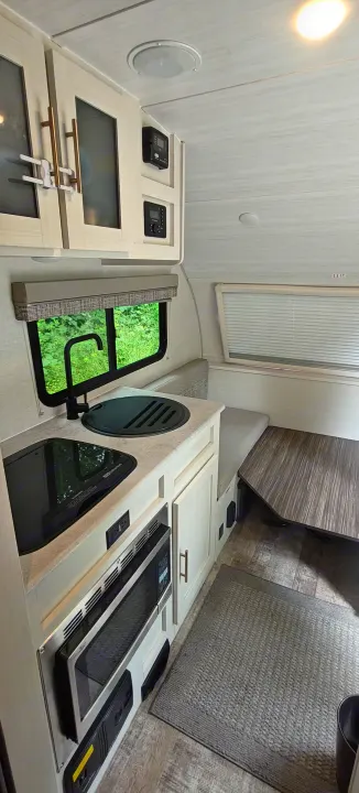 Modern RV rental interior: kitchen with sink, stovetop, microwave, and dinette seating by a window with green view.. Forest River R-Pod 2023