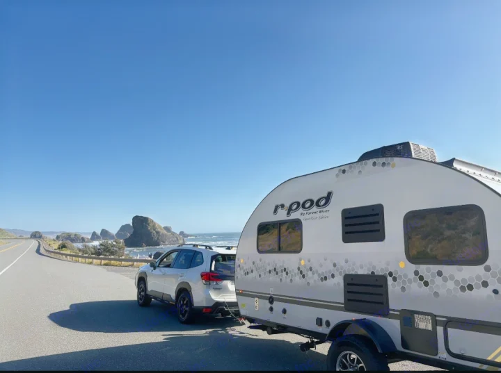 Enjoying a scenic coastal road trip with our RV rental. A white SUV tows an r-pod travel trailer next to the ocean and sea stacks.. Forest River R-Pod 2023